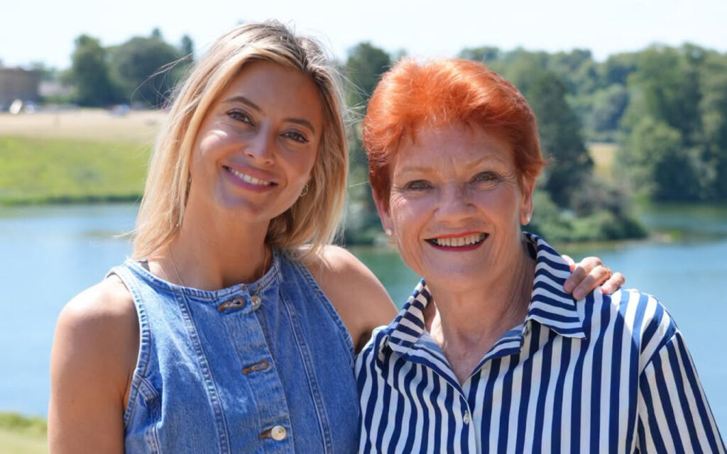 Two women smiling at the camera by a lake, with one in a denim sleeveless top and the other in a striped shirt, arm around each other.