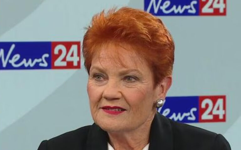 Close-up of a woman with short red hair in a black blazer, speaking in a News 24 TV studio backdrop.