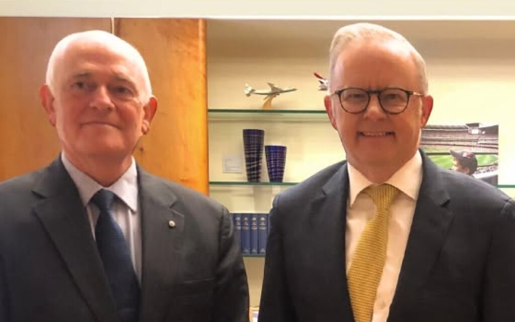 Two older men in suits posing for a photo in an office, one with a navy tie and the other with a yellow tie.