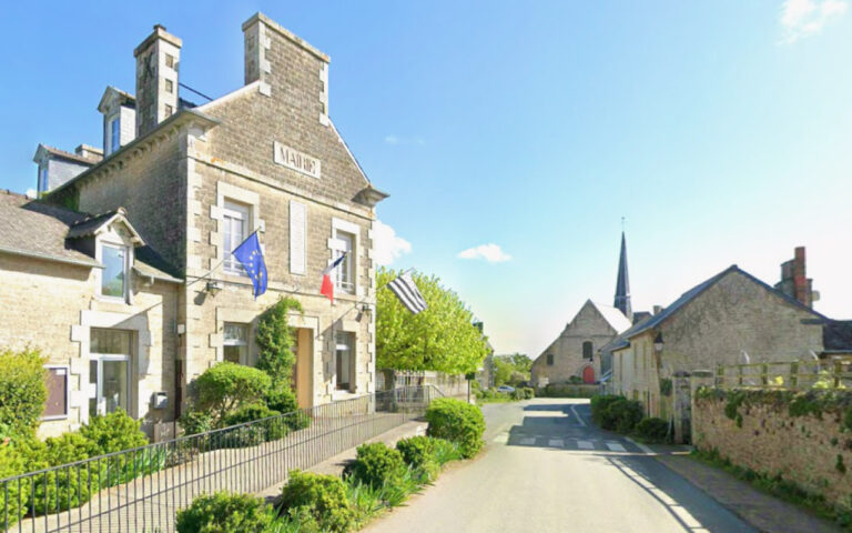 Stone town hall (mairie) with EU and French flags on a sunny village street; a church spire visible in the distance.