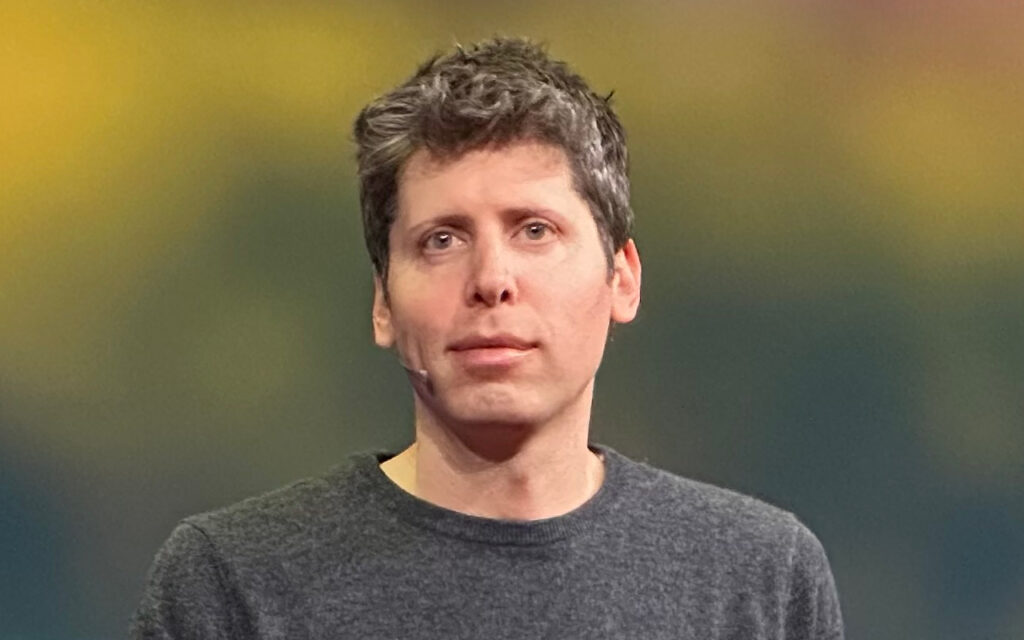 Speaker wearing a lavalier microphone on stage, facing the audience with a gray sweater and blurred backdrop.