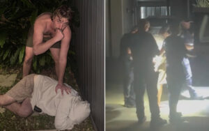 Shirtless man leans over a person lying facedown on the ground beside a wall, appearing threatening outdoors.