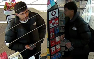 Two men in a store near a checkout; one wearing a black puffer jacket and mask under the chin holds a small item, the other at a kiosk holding a bottle and backpack.