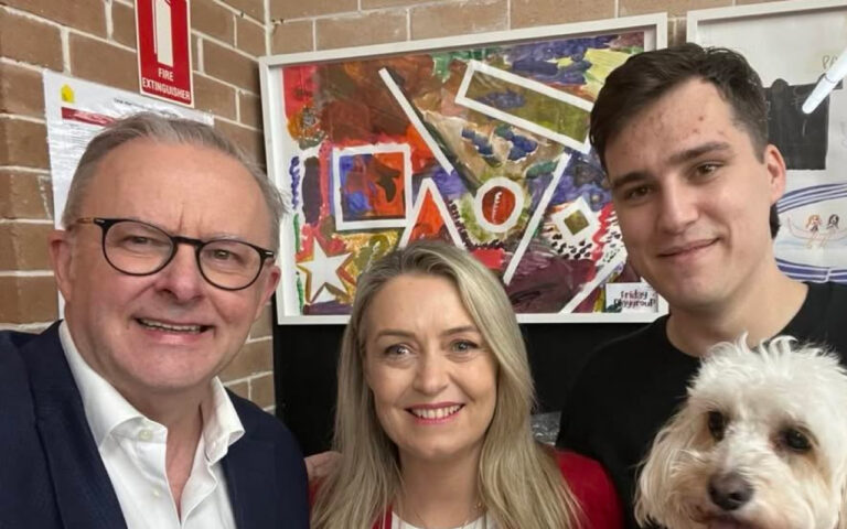 Three people smile for a photo with a fluffy white dog in front of a bright abstract painting on the wall.