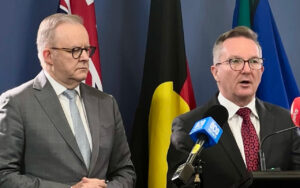 Two men in suits at a press conference with multiple flags behind them and microphones in front of the speaker on the right.