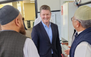 Smiling man in a blue suit talks with two older men wearing skullcaps inside a shop or showroom.