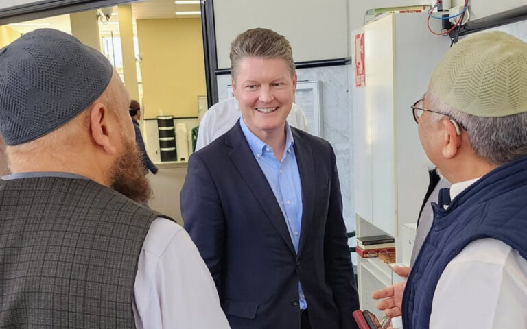 Smiling man in a blue suit talks with two older men wearing skullcaps inside a shop or showroom.