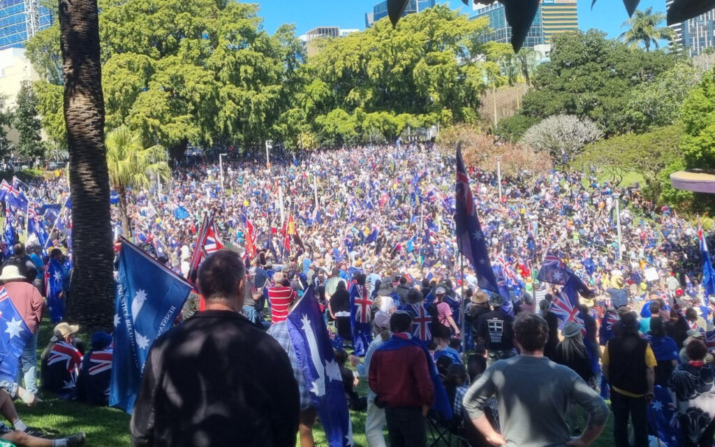 Large crowd gathered in a sunny city park, many waving Australian flags and blue banners, with trees and buildings in the background.