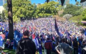 Large crowd gathered in a sunny city park, many waving Australian flags and blue banners, with trees and buildings in the background.