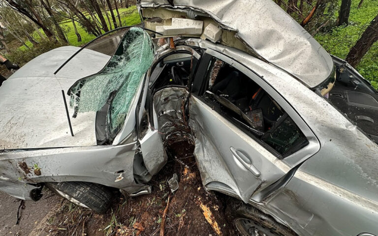 Silver sedan severely damaged in a roadside crash, with a shattered windshield and open front doors among trees and mud.