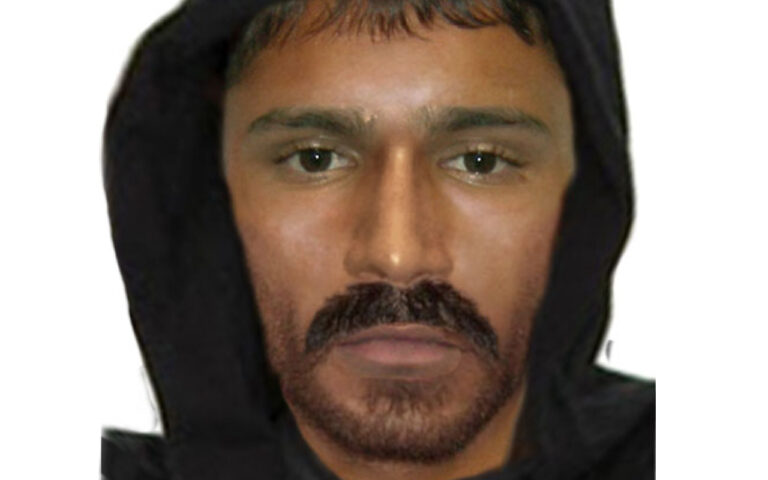 Man wearing a black hood, with short dark hair and a trimmed mustache and beard, looking at the camera.