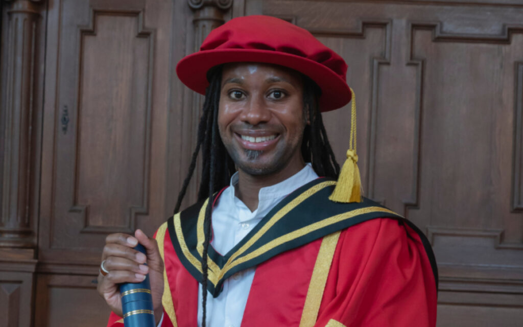 Smiling graduate in a red gown and hat with black and gold hood, holding a diploma tube in front of a wood-paneled background.