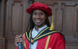 Smiling graduate in a red gown and hat with black and gold hood, holding a diploma tube in front of a wood-paneled background.