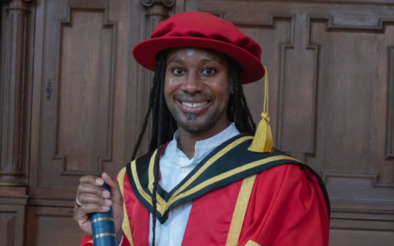 Smiling graduate in a red gown and hat with black and gold hood, holding a diploma tube in front of a wood-paneled background.