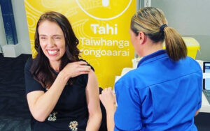 Smiling woman receives a vaccine injection in her upper arm from a nurse in a blue uniform, with a bright yellow banner in the background.