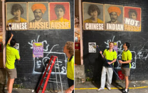 Two workers in bright shirts paint graffiti on a black brick wall beneath large vintage portrait posters; a red ladder leans against the wall.