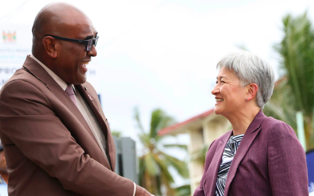 Two professionals smile and shake hands outdoors, palm trees in the background, signifying a friendly meeting.