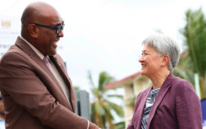 Two professionals smile and shake hands outdoors, palm trees in the background, signifying a friendly meeting.