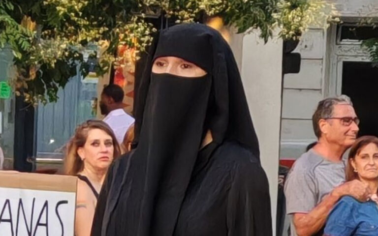 A person wearing a black niqab and abaya standing in a street with pedestrians in the background (face mostly covered).