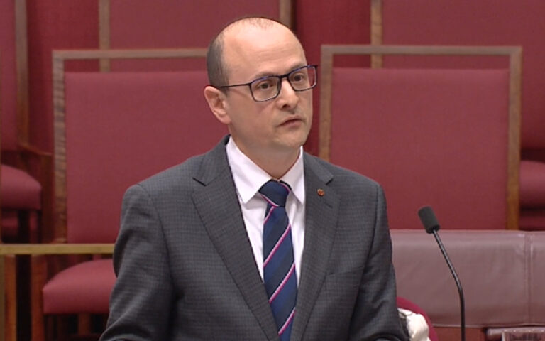 A man in a gray suit and striped blue tie speaks at a podium in a formal chamber with pink seating in the background.