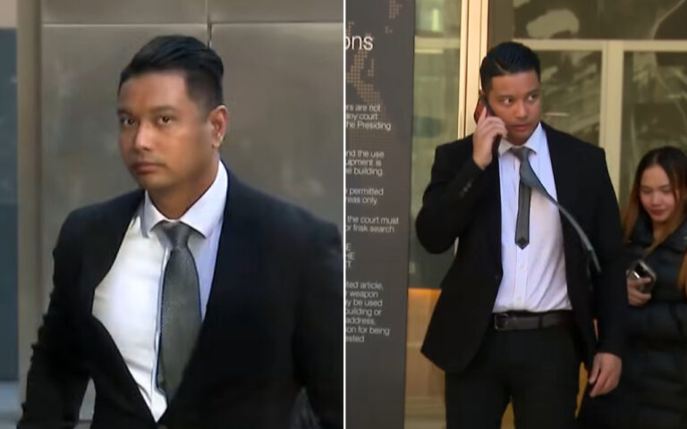 A man in a dark suit and gray tie walking toward the camera outside a building, looking straight ahead.