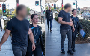 Man in a dark T‑shirt and jeans walks beside a female staff member with a badge along a sunny urban sidewalk near a crosswalk and traffic lights.