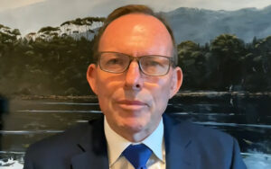 Man in a dark suit and blue tie speaks to the camera against a mural of a forest by a lake, wearing glasses and a neutral expression.