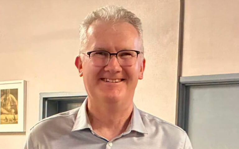 Smiling man with short gray hair and glasses, wearing a light gray button-down shirt indoors.