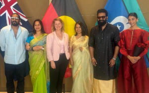 Group of six adults posing for a photo at a formal event, standing in front of international flags, dressed in colorful attire including sarees and traditional outfits.