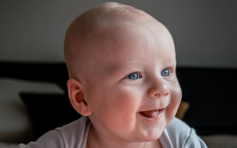 Smiling baby with blue eyes looking to the right, wearing a light gray outfit.