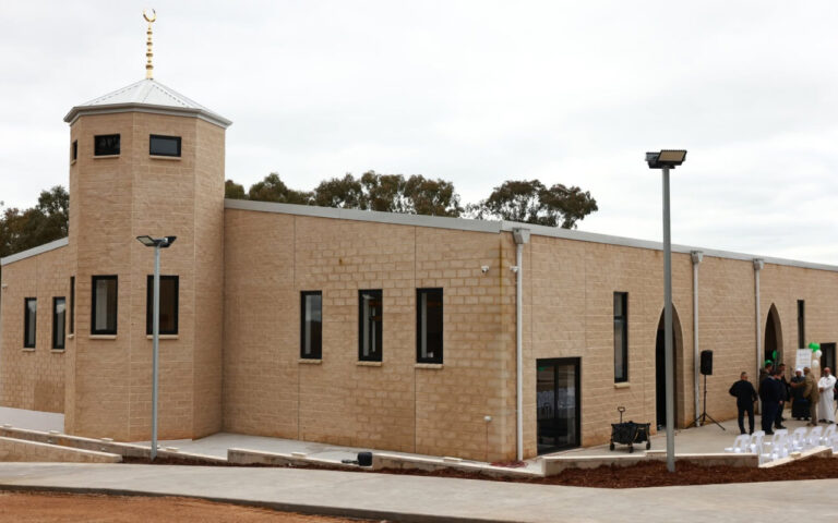 Beige brick building with a small tower and arched entrance; group of people gathering near the doorway and white chairs outside.